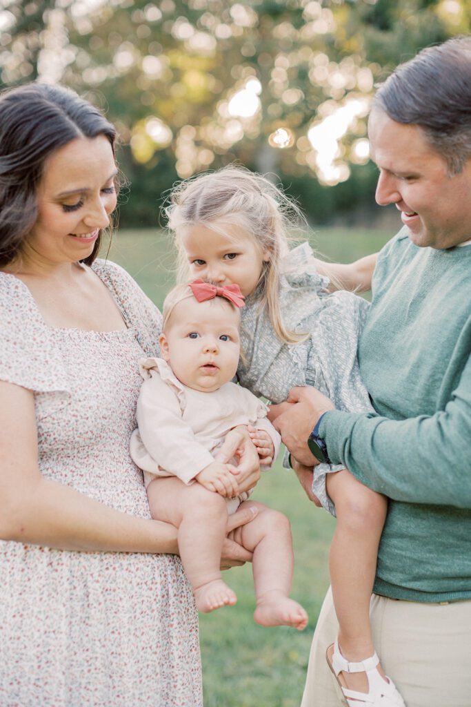 Little blonde girl leans down to kiss her baby sister's head during their family photo session.