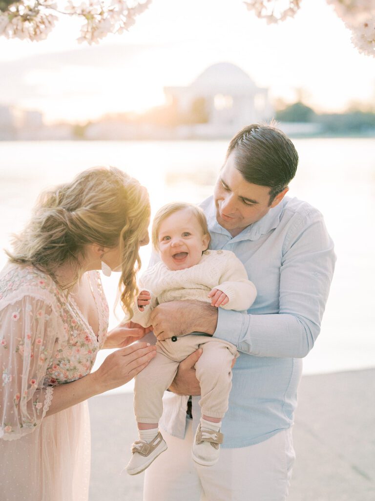 Parents Hold Infant Son And Tickle Him To Make Him Laugh With A View Of The Jefferson Memorial In The Background.