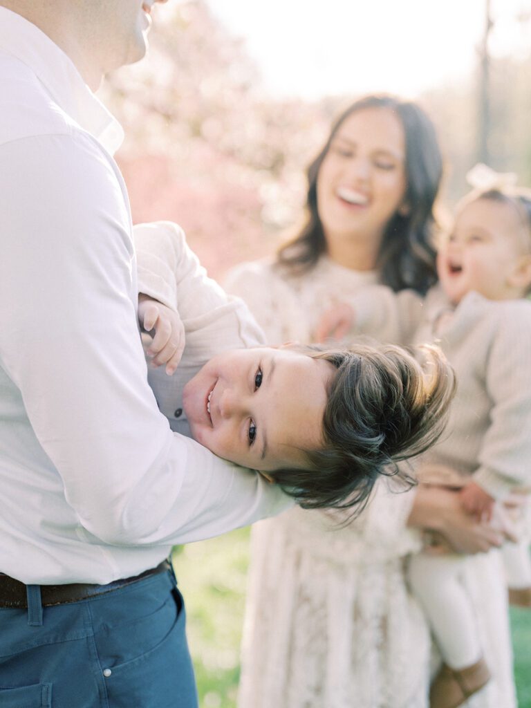 Little Boy Smiles At The Camera While His Father Swings Him Around While Mother Holds Toddler Daughter.