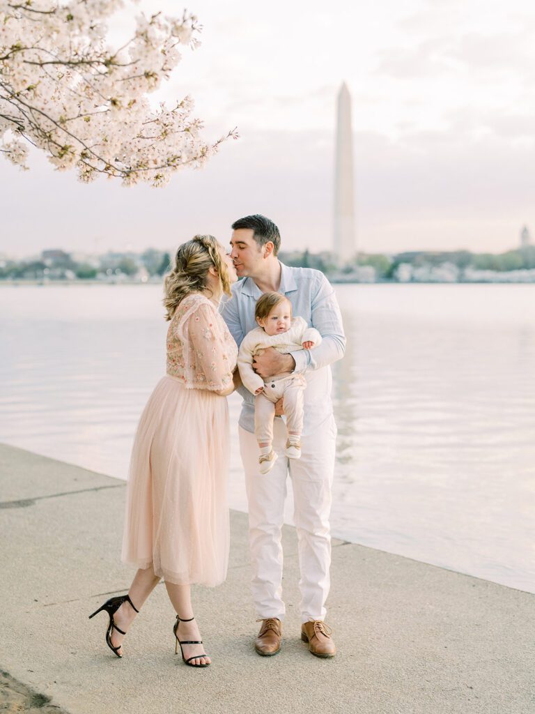 Mother And Father Lean In For A Kiss While Holding Their Son During Their Dc Cherry Blossoms Family Session.