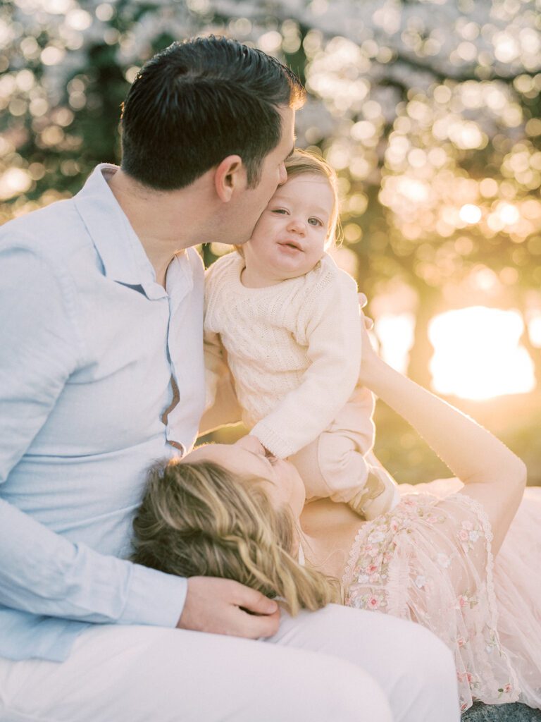 Mother Lays In Husband's Lap While Holding Up Her Infant Son For A Kiss From Her Husband.