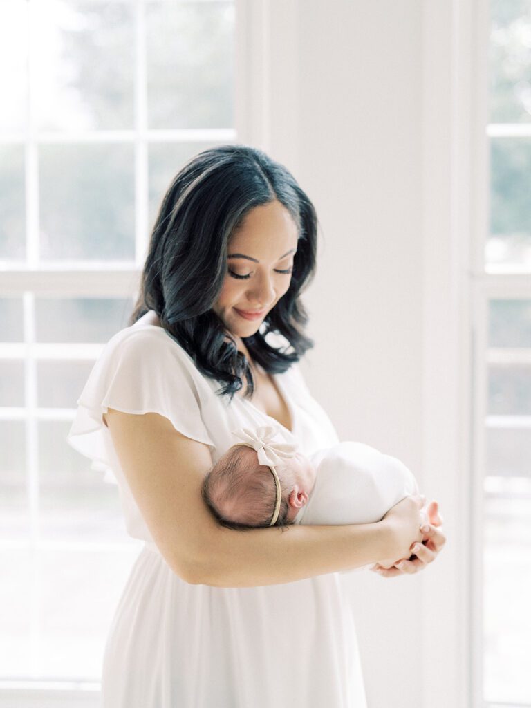 A Mother With Dark Hair In A White Dress Smiles Down At Her Newborn Baby.