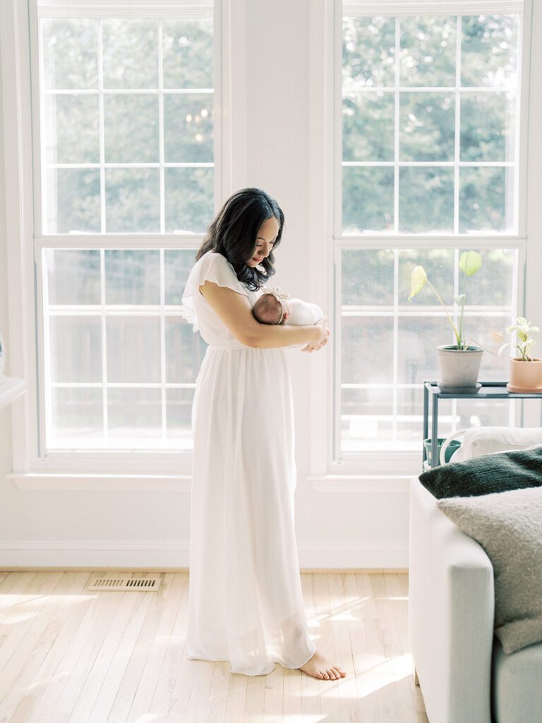 Mother With Dark Hair In A White Dress Walks Her Newborn Baby In Her Home.