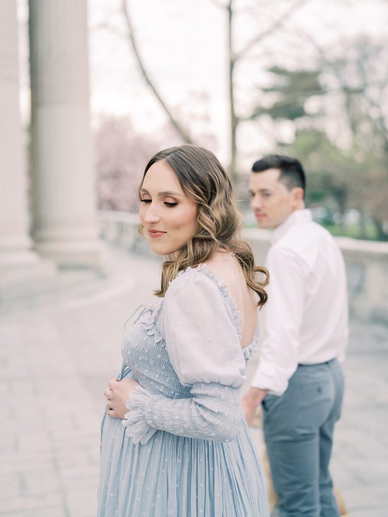 Pregnant Mother In Blue Doen Dress Places On Hand On Top Of Her Belly While Her Husband Looks Back At Her During Their Maternity Session At The Dar Constitution Hall.