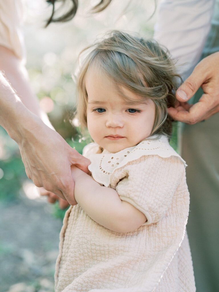 A toddler daughter stands looking at the ground while parents hold her hands and touch her head.