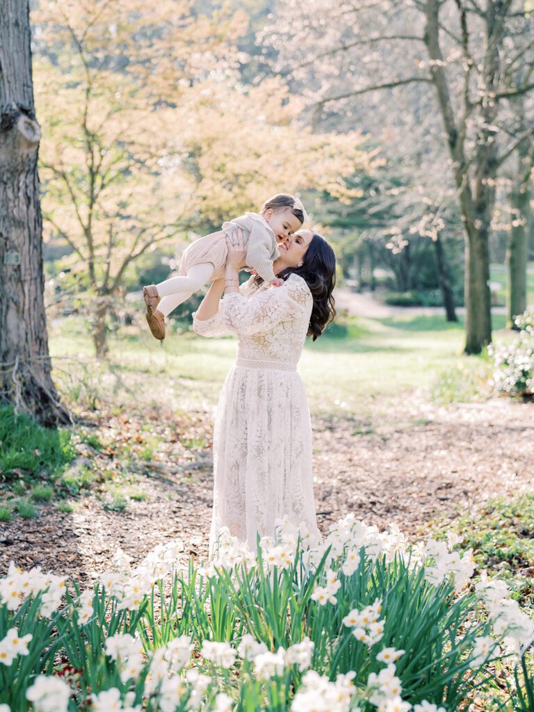 Mother In White Dress Holds Up Her Toddler Daughter In The Daffodils At Brookside Gardens In Silver Spring, Maryland.