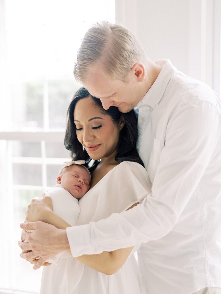 Blonde Father Leans Into His Dark-Haired Wife Who Holds Their Newborn Baby.