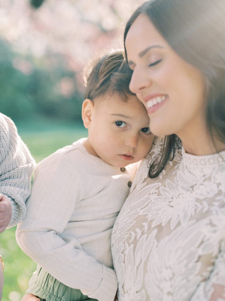 Little Boy Looks At The Camera While Mother Holds Him With Eyes Closed.