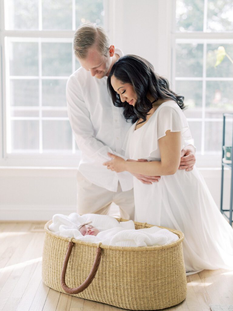 Interracial Couple Kneels Next To A Moses Basket By A Large Window During Their Newborn Session.