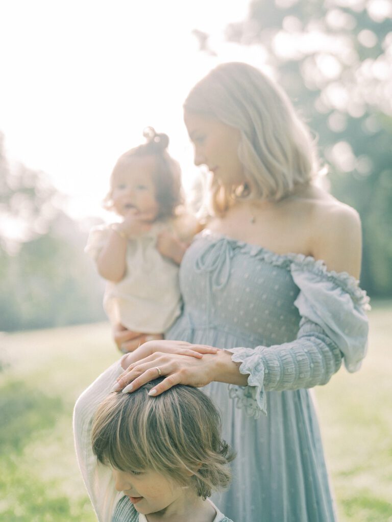 A Blonde Mother Stands In A Field With Her Hand On Her Son's Head.