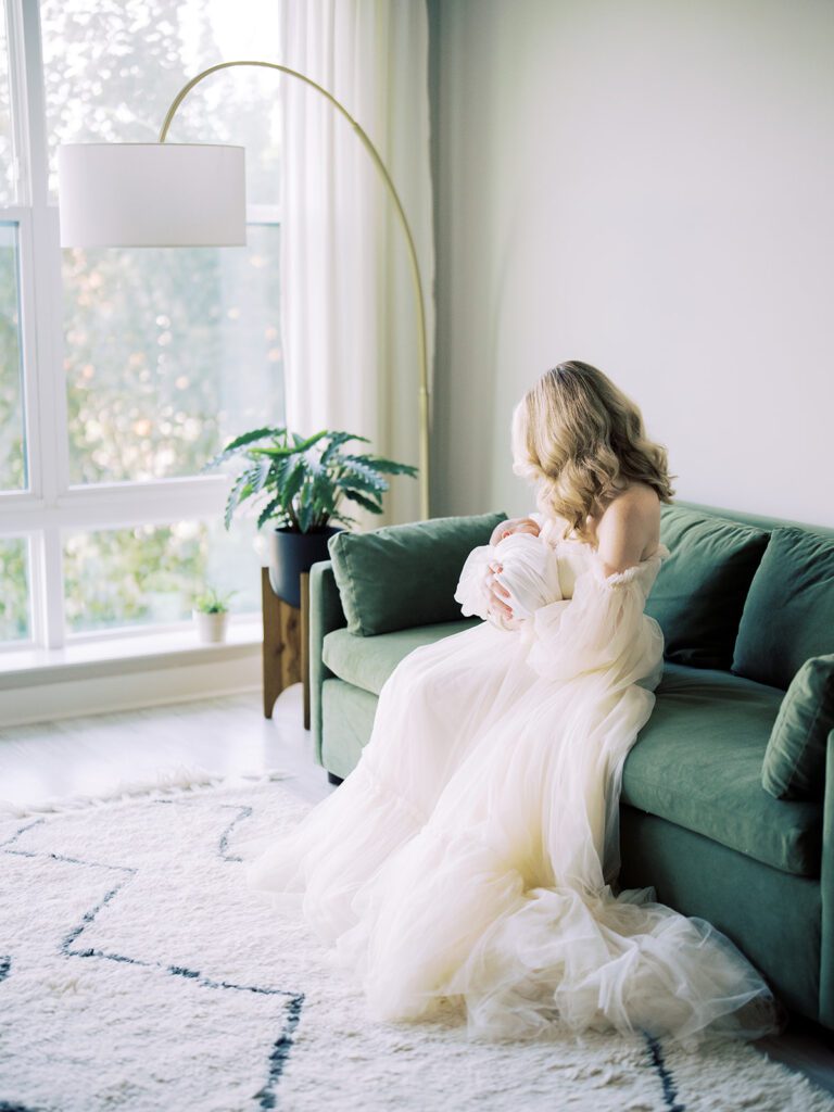 Blonde Mother Sits On A Green Velvet Couch Looking Down At Her Newborn Son.