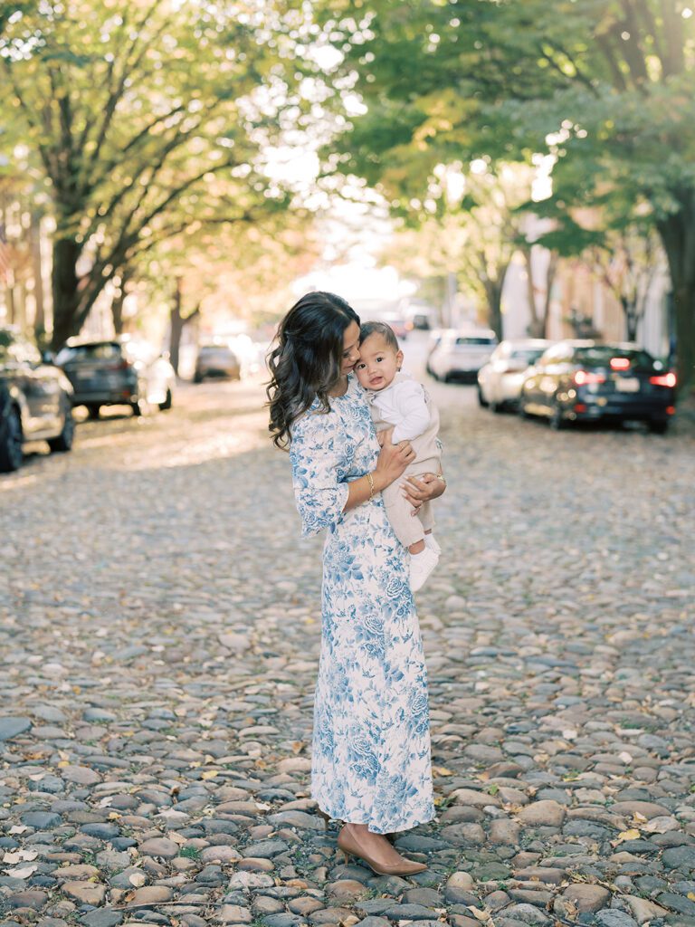 A mother in a blue floral dress holds her young son while standing on Prince Street in Alexandria, Virginia.