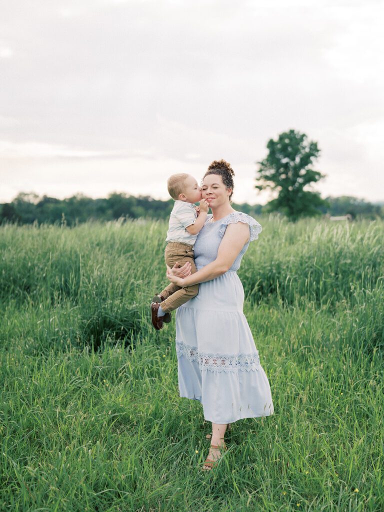 Little Boy Kisses His Mother's Cheek As She Stands In A Green Field, Photographed By Manassas Photographer Marie Elizabeth Photography.