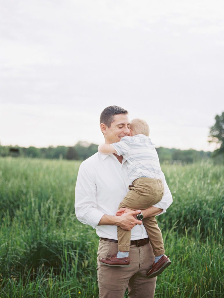 Father Holds His Son While Standing In A Green Field.