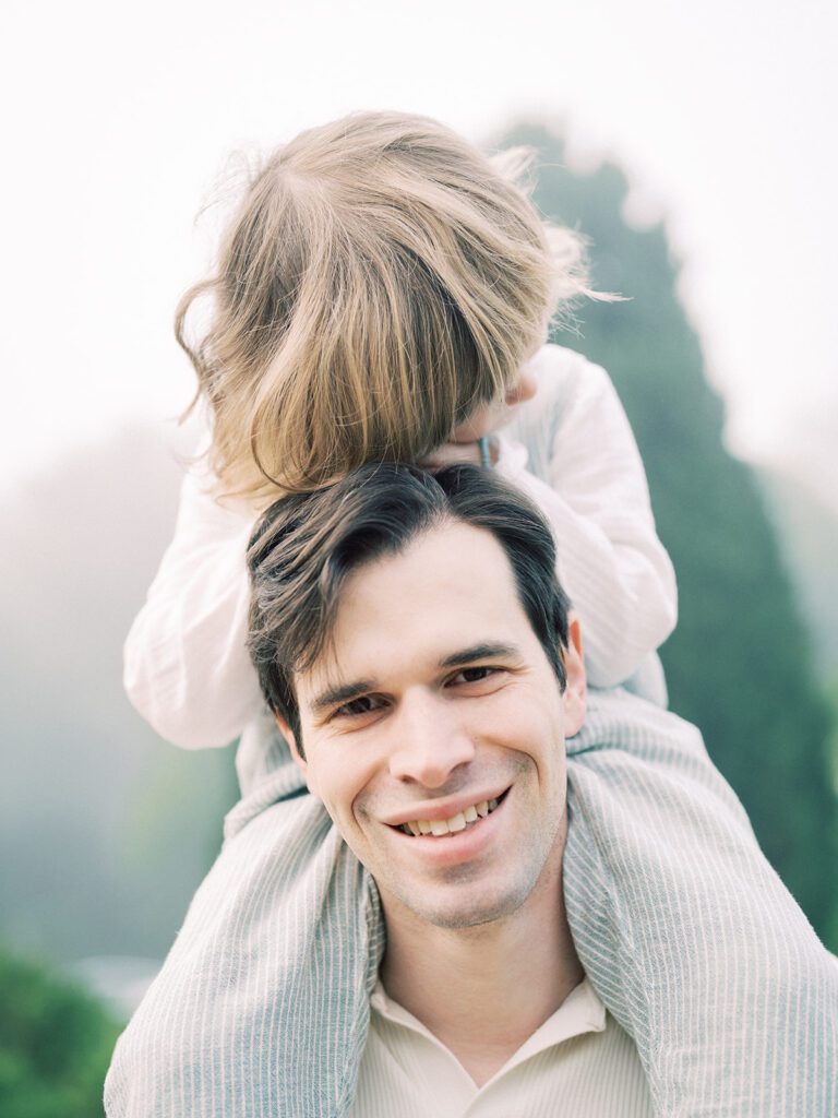 Father Smiles At The Camera As His Son Sits On His Shoulders.