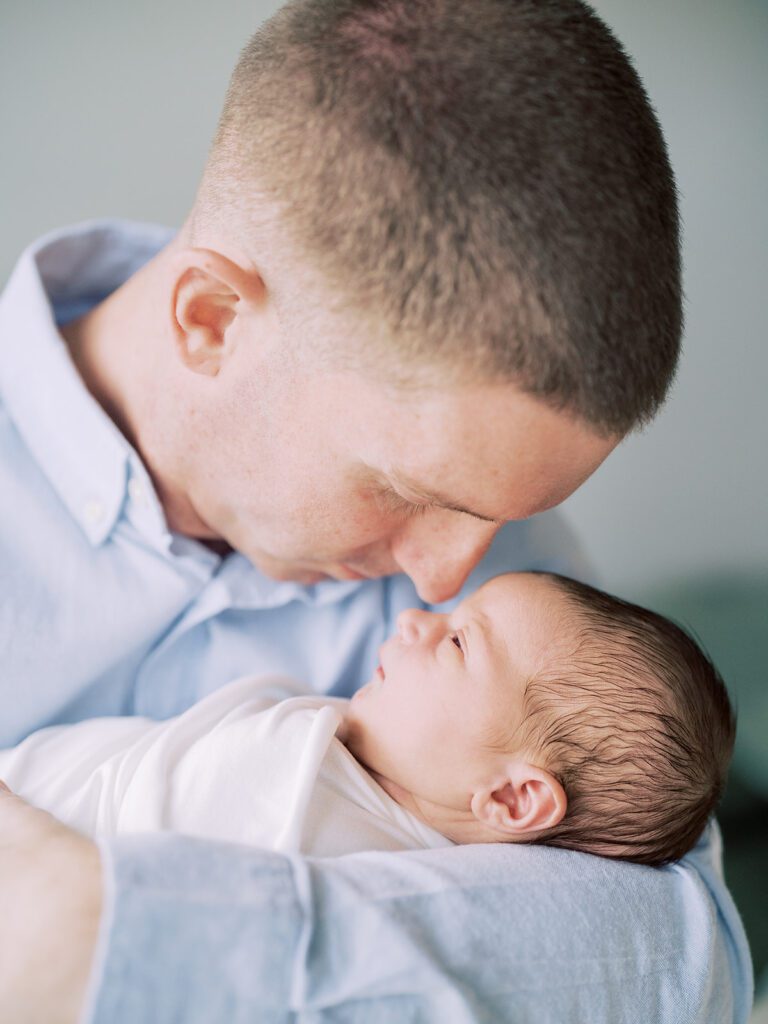 A Dad In A Long-Sleeved Blue Shirt Holds His Newborn Baby Up Nose-To-Nose.