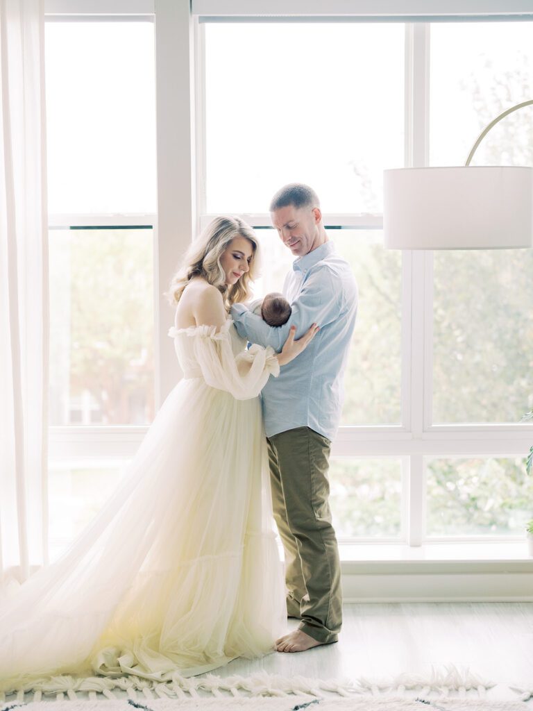 A Blonde Mother In A Long Cream Gown Stands With Her Husband In Front Of A Large Window While They Hold Their Newborn Son.