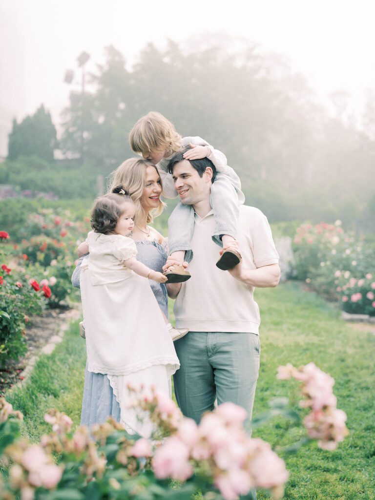 Family Of Four Stands In The Roses At Bishop's Garden During Their Family Photo Session.