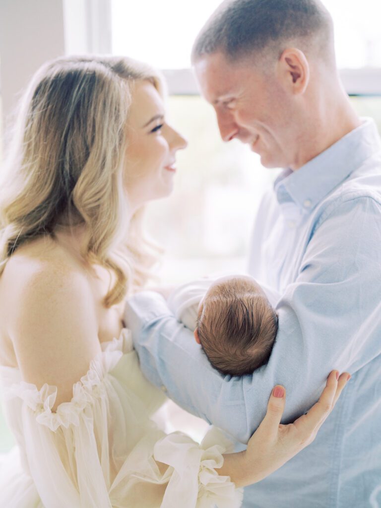 New Parents Stand In Front Of A Window Holding Their New Baby Smiling At One Another.