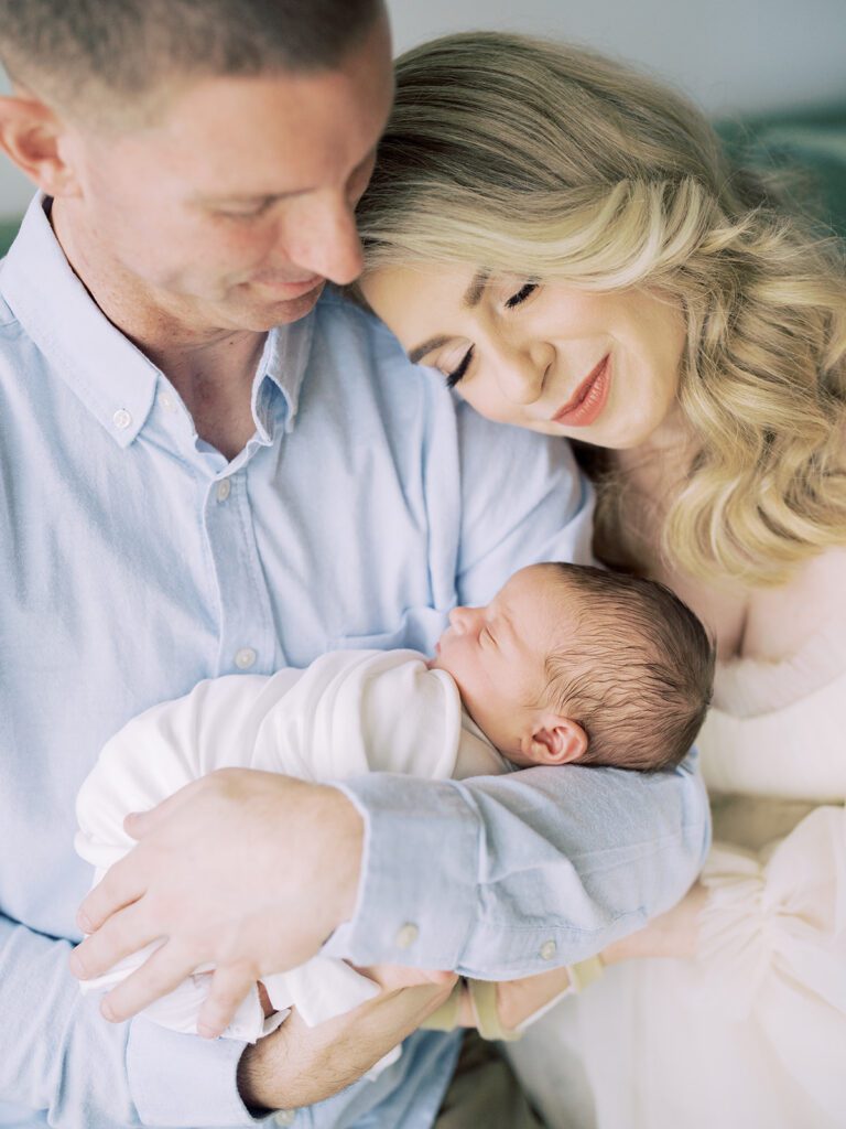 A Blonde Mother Rests Her Head On Her Husband's Shoulder While He Holds Their Newborn Baby.