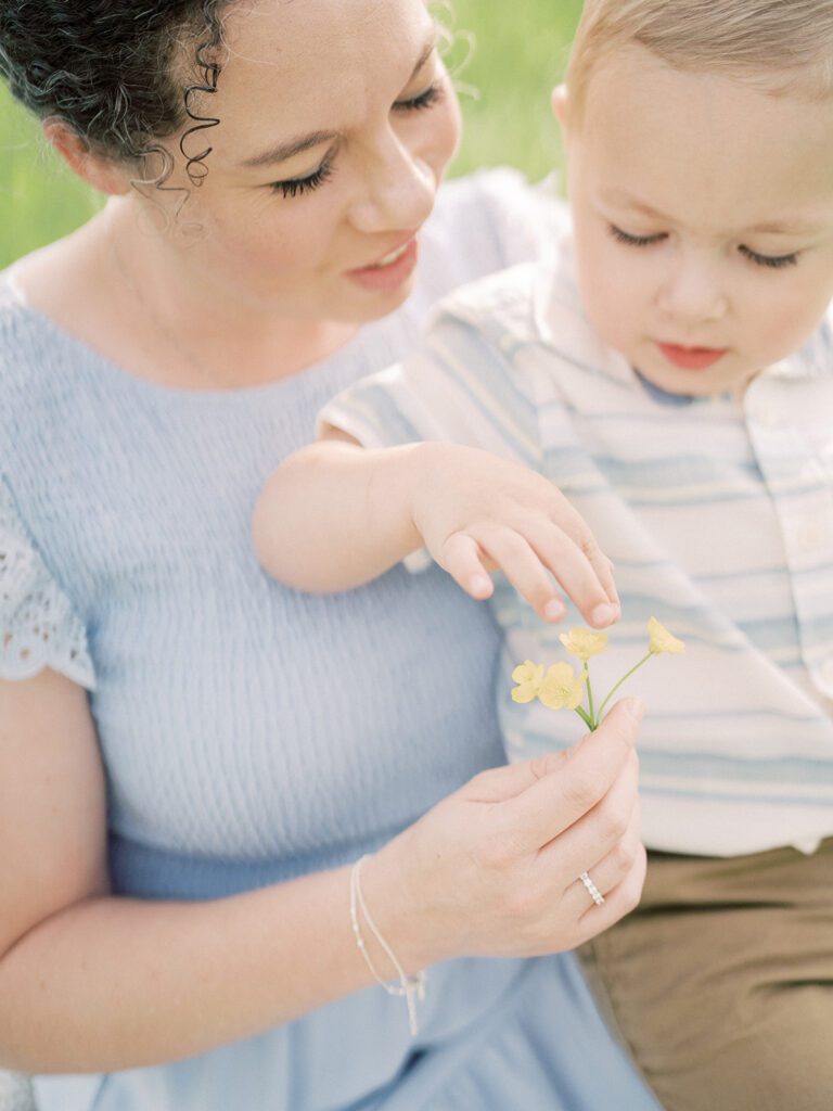 Little Boy Sits On His Mother's Lap And Touches Flowers In Her Hand.