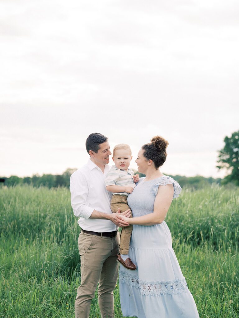 Mother And Father Stand Together With Their Young Son In A Field.