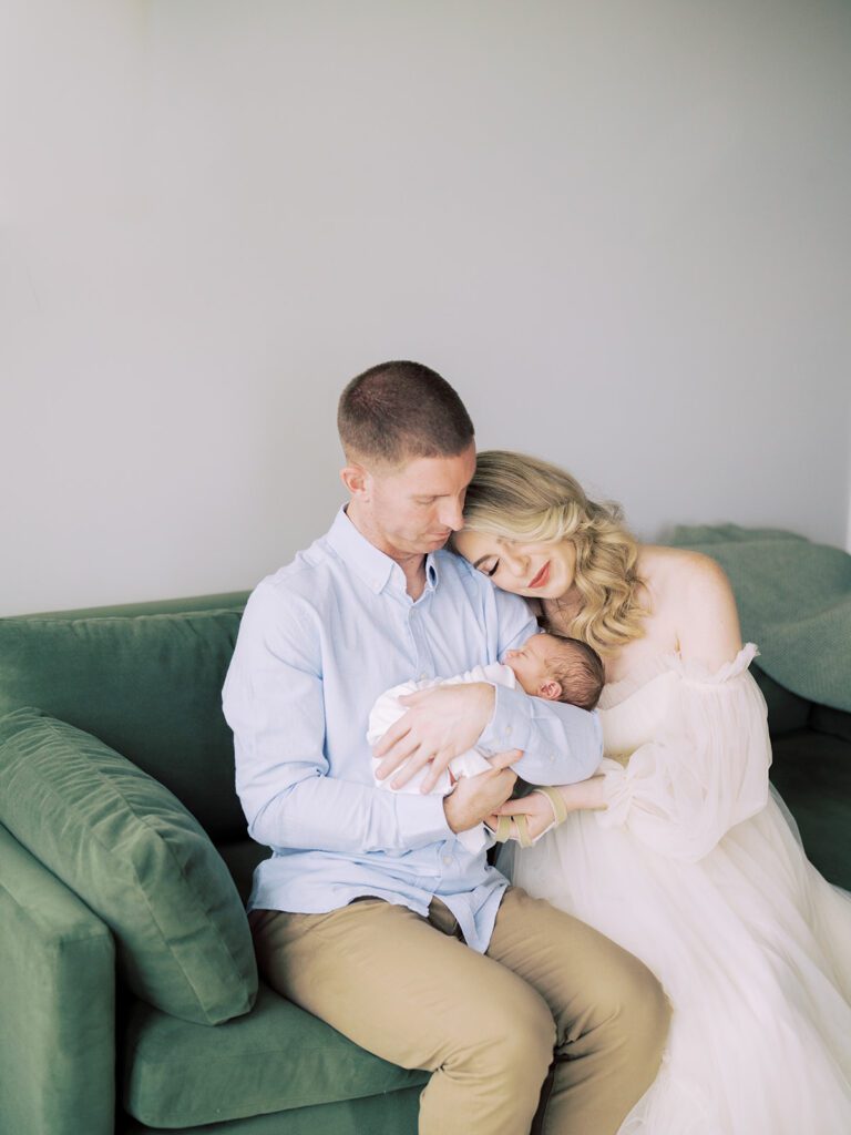 A Blonde Mother In A Cream Gown Rests Her Head On Her Husband's Shoulders While He Holds Their Newborn Son During Their Newborn Session.