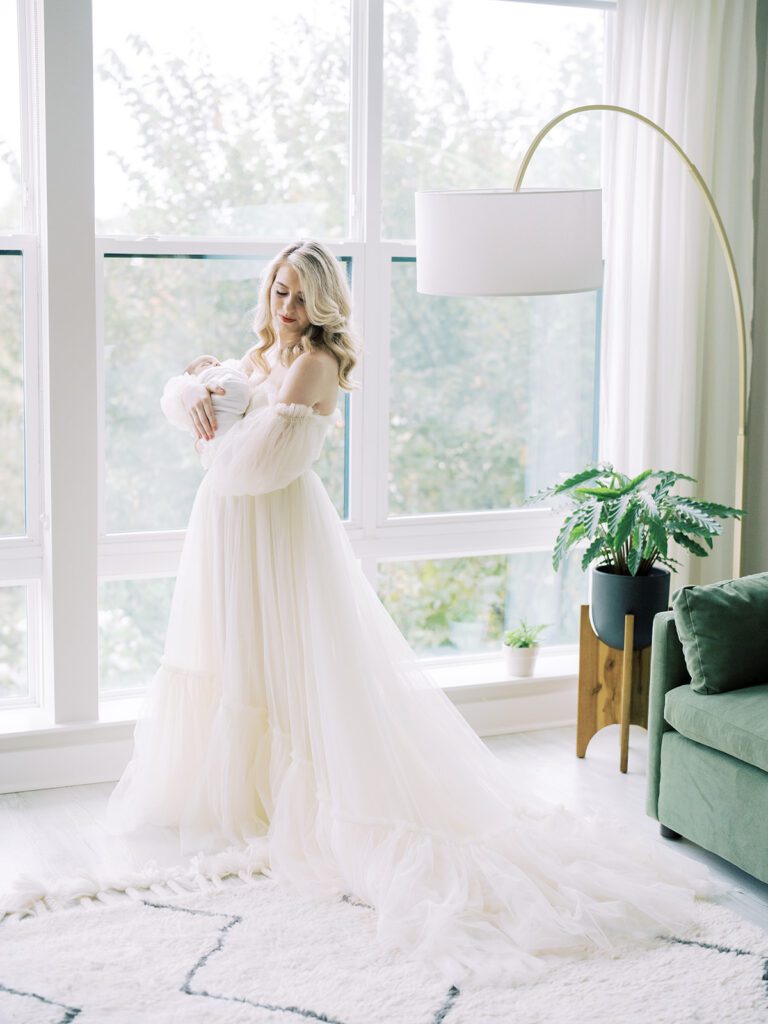 A Blonde Mother Wearing A Long Cream Gown Holds Her Newborn Son While Standing New A Large Window In Her Living Room.