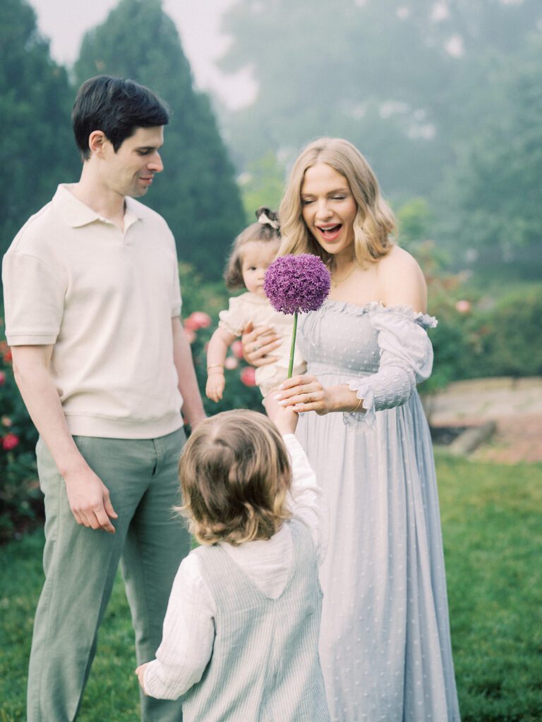 A Big Brother Brings A Purple Flower Over To His Mother As She Stands Holding Her Daughter With Her Husband.