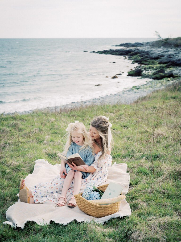 A Mother Sits With Her Young Daughter On Her Lap Reading A Book Along The Rhode Island Coast.