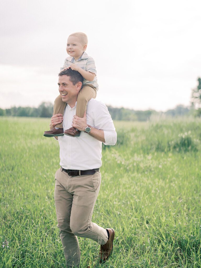 A Father Runs Through A Field With His Toddler Son On His Shoulders.