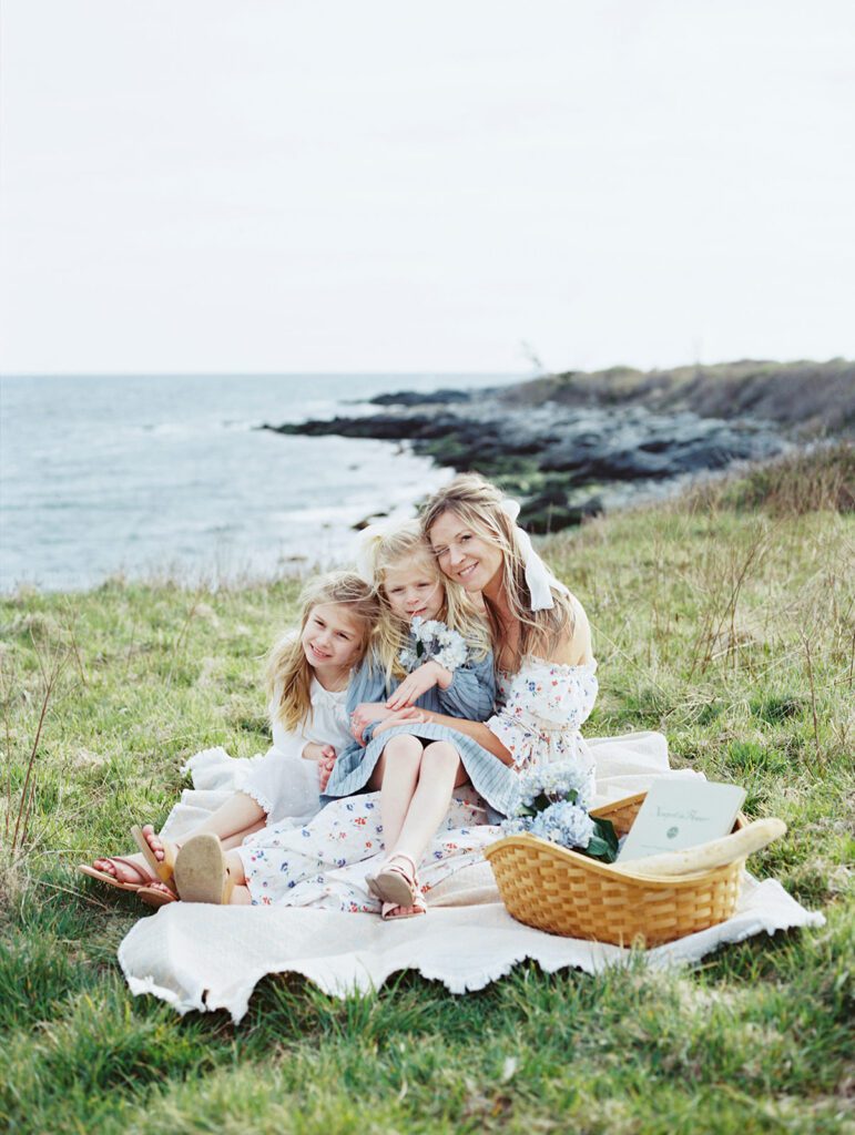 A Mother Sits With Her Two Daughters In Grass Along The Coast, Smiling At The Camera.