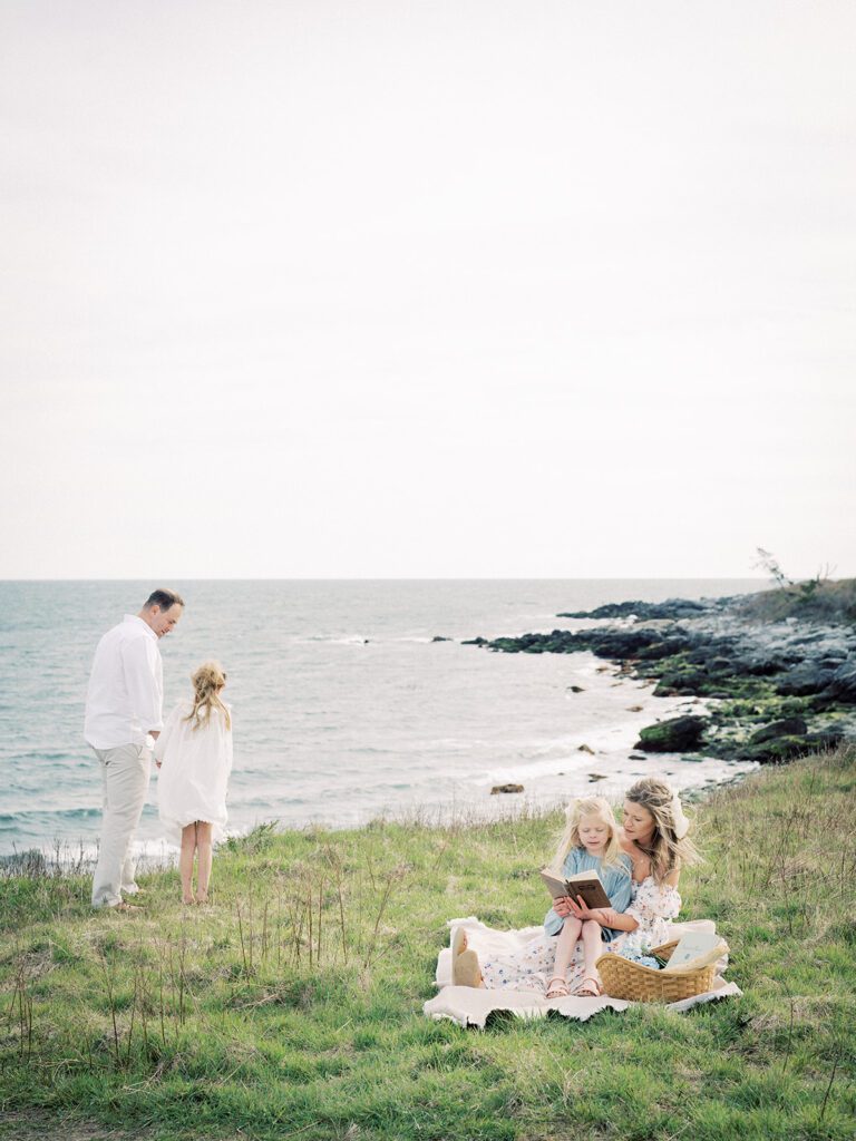 A Mother Sits With Her Daughter On Her Lap As Her Husband Walks With Their Other Daughter Along A Luscious Coastline, Photographed By Destination Family Photographer, Marie Elizabeth Photography.