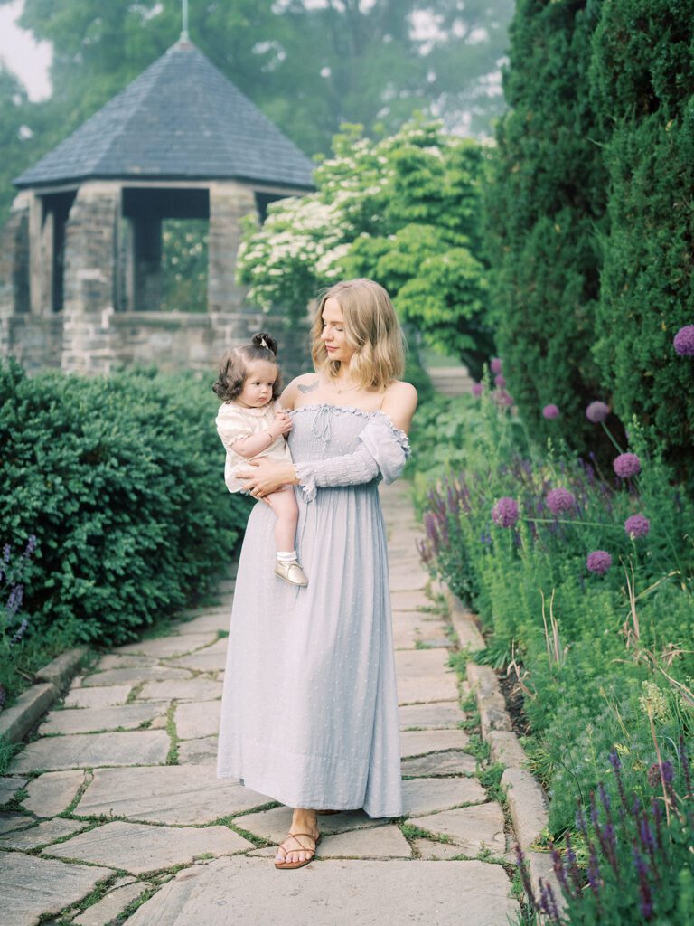 Blonde Mother Stands On Stone Path With Her Young Daughter During Her Family Photo Session At Bishop's Garden In Dc.