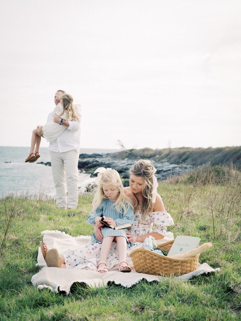 Mother Sits With Her Daughter On Her Lap As Her Husband Picks Up And Swings Their Other Daughter As They Sit Along A Beach, Photographed By Destination Family Photographer, Marie Elizabeth Photography.