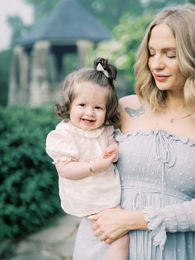 Blonde Mother Holds Her Baby Girl In A Garden.