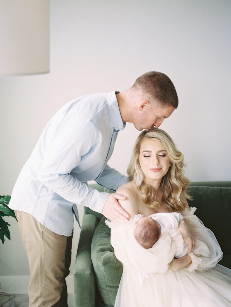 A Father In A Long-Sleeved Blue Shirt Leans Over A Couch To Kiss His Blonde Wife Who Holds Their Newborn Son, Photographed By Bethesda Newborn Photographer Marie Elizabeth Photography.