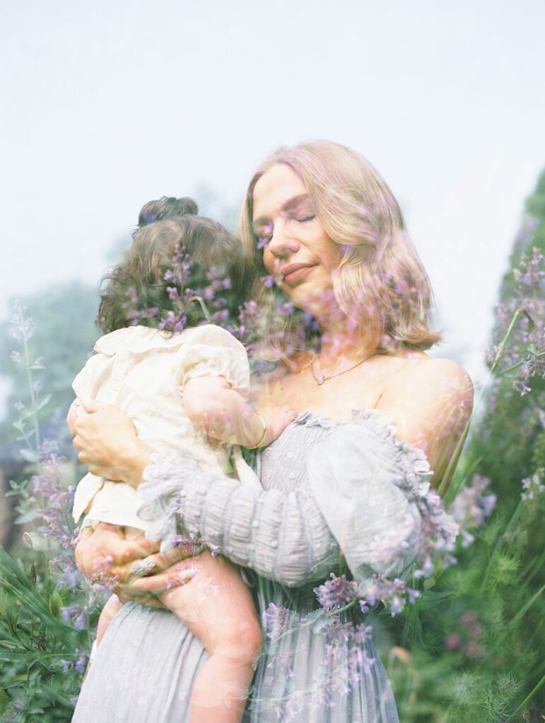 A Double Exposure Of A Mother Holding Her Daughter Against Purple Blossoms.