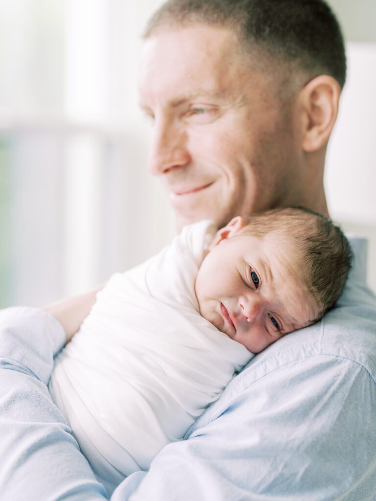 A Newborn Baby Rests On His Father's Chest As His Father Looks Off Smiling.