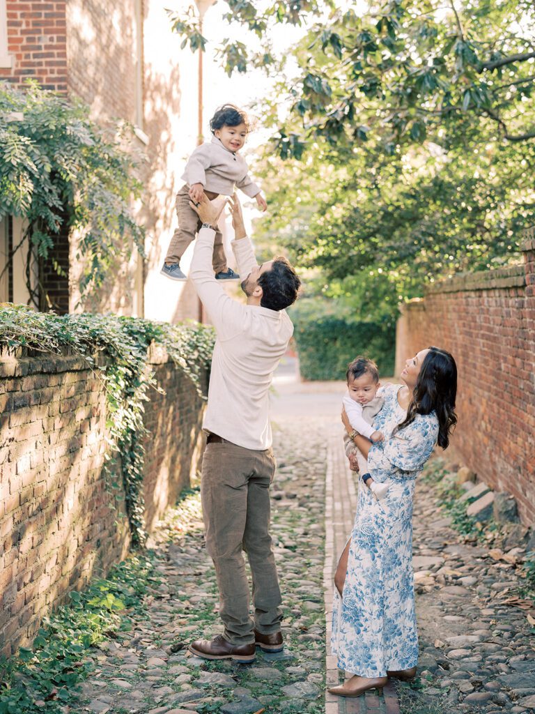 A dad throws his son up in the air as he stands in an alley with his wife.