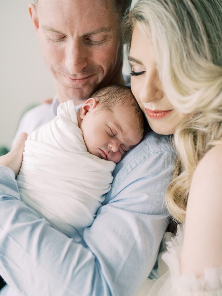 A Blonde Mother Leans Into Her Husband Wearing A Blue Shirt While Holding Their Swaddled Newborn Son, Photographed By Bethesda Newborn Photographer Marie Elizabeth Photography.