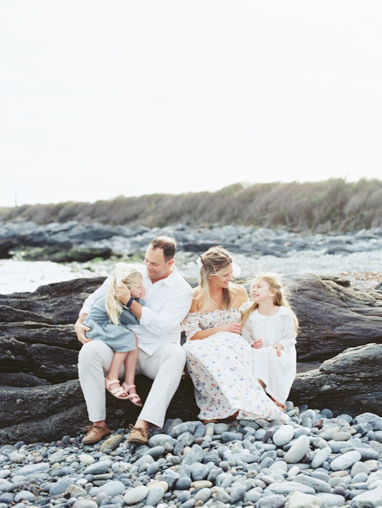 Family Of Four Sits On A Log Along A Rocky Beach, Photographed By Destination Family Photographer, Marie Elizabeth Photography.