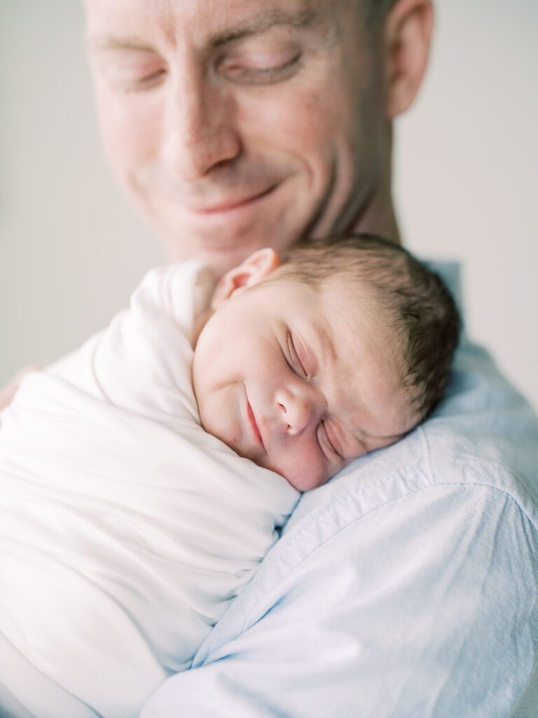 A Father Smiles At His Newborn Son As His Newborn Son Gives A Soft Smile While Sleeping On His Chest.