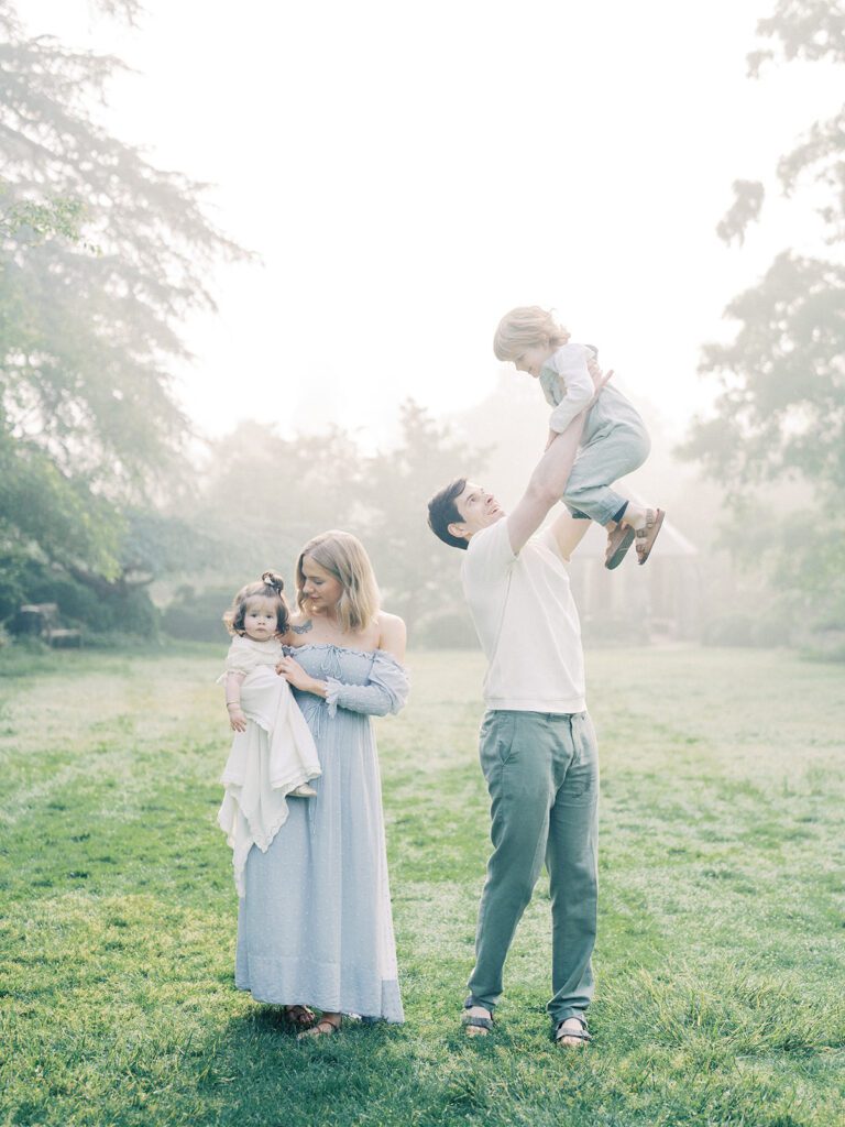 A Father Throws His Son Up In The Air While A Mother Stands Holding Her Daughter.