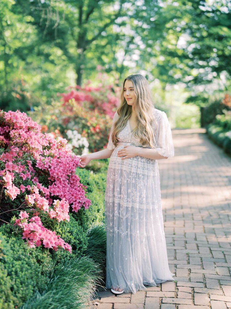 Pregnant Mom In Purple Needle &Amp; Thread Gown Stands In A Garden With One Hand On A Pink Azalea Bush During Her National Arboretum Azaleas Maternity Session.