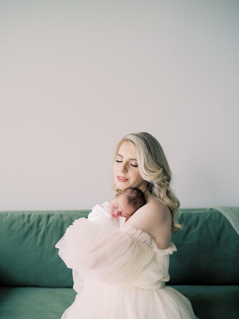 A Blonde Mother In A Cream Gown Sits On A Green Velvet Couch Holding Her Newborn Son, Photographed By Bethesda Newborn Photographer Marie Elizabeth Photography.