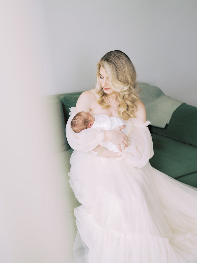 Blonde Mother In Cream Dress Sits On Green Couch Holding Newborn Son.