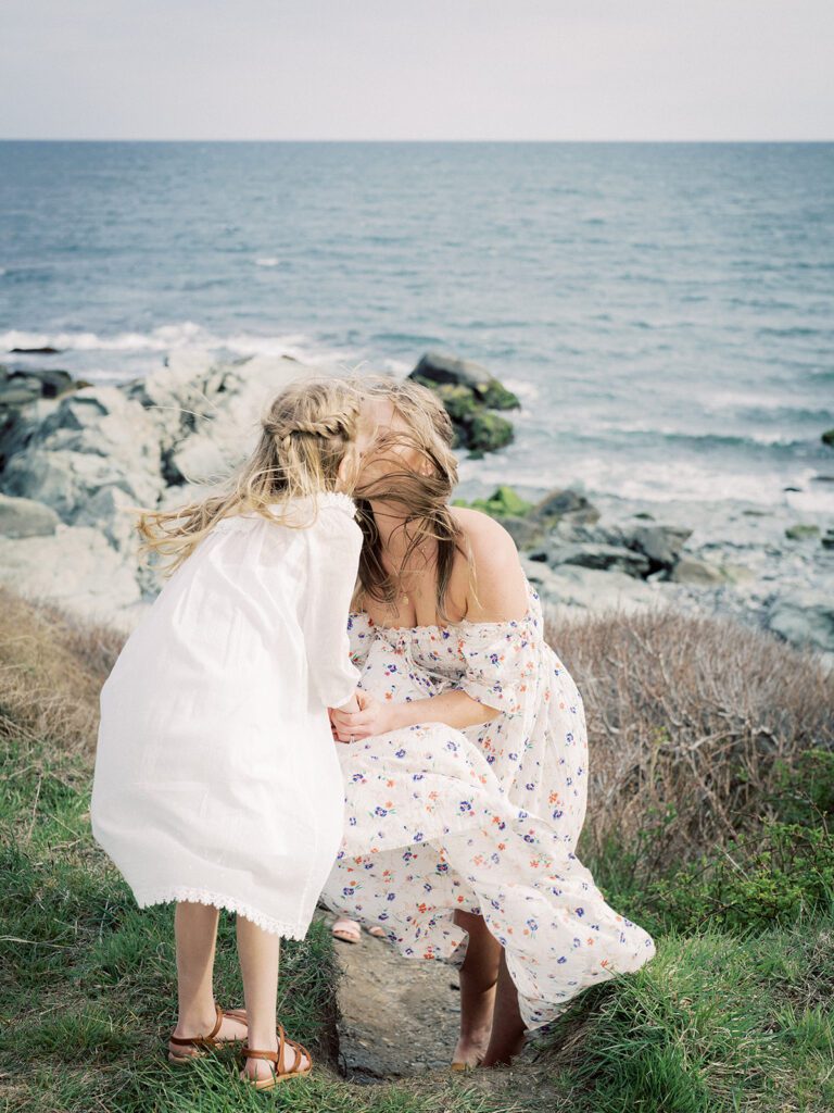 A Little Girl And Her Mother Lean In For A Kiss As The Wind Blows Their Hair Along The Grassy Plains Near The Beach.