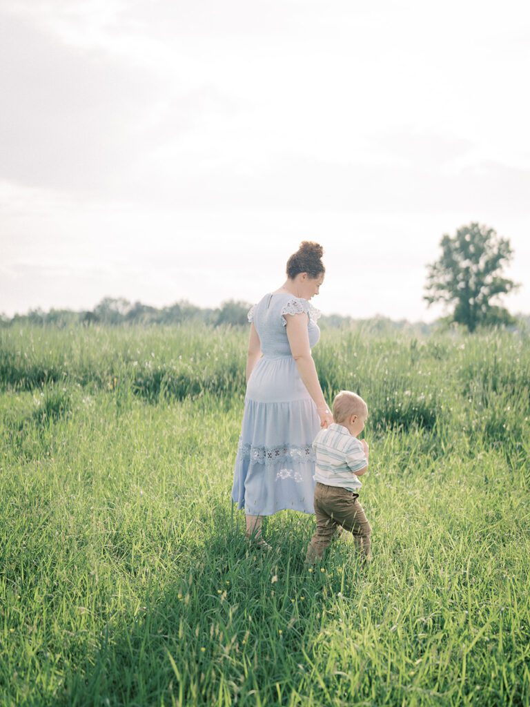 Mother In Blue Dress Walking Through A Field With Her Toddler Son, Photographed By Manassas Photographer Marie Elizabeth Photography.