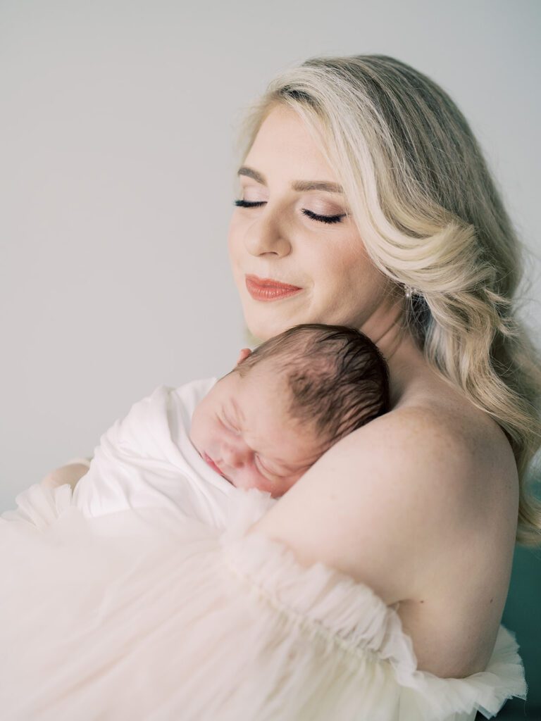 A Blonde Mother Holds Her Newborn Baby Swaddle In White On Her Chest And Closes Her Eyes, Photographed By Bethesda Newborn Photographer Marie Elizabeth Photography.